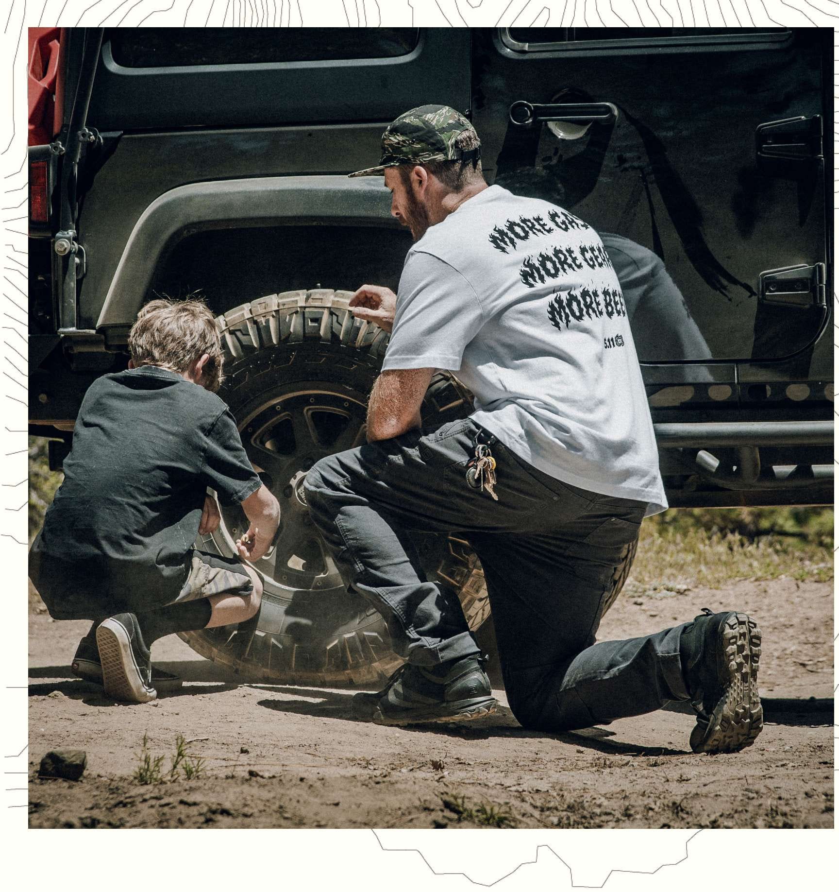 A man and child kneel beside the wheel well of an off-road vehicle.