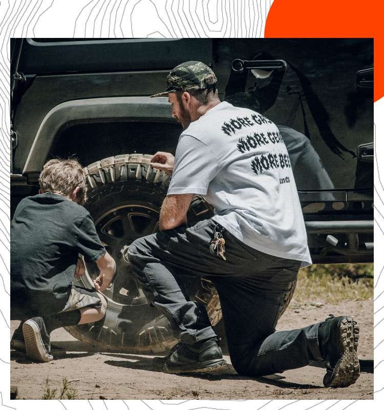 A man and child kneel beside the wheel well of an off-road vehicle.