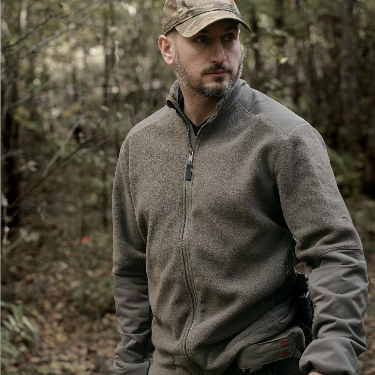 Hombre en el bosque con un forro polar táctico verde ranger y una gorra de camuflaje.