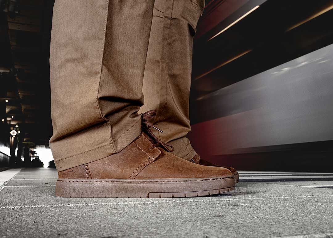 Jemand steht auf einem Bahnsteig, während ein Zug vorbeifährt. Die Person trägt braune, moderne Chukka-Stiefel.