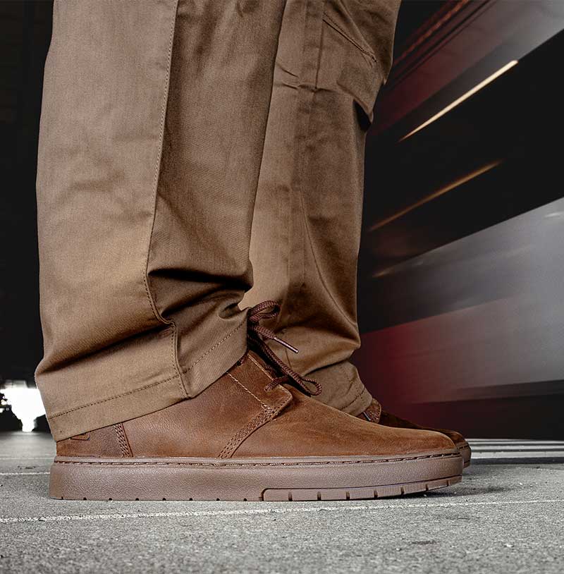 Jemand steht auf einem Bahnsteig, während ein Zug vorbeifährt. Die Person trägt braune, moderne Chukka-Stiefel.