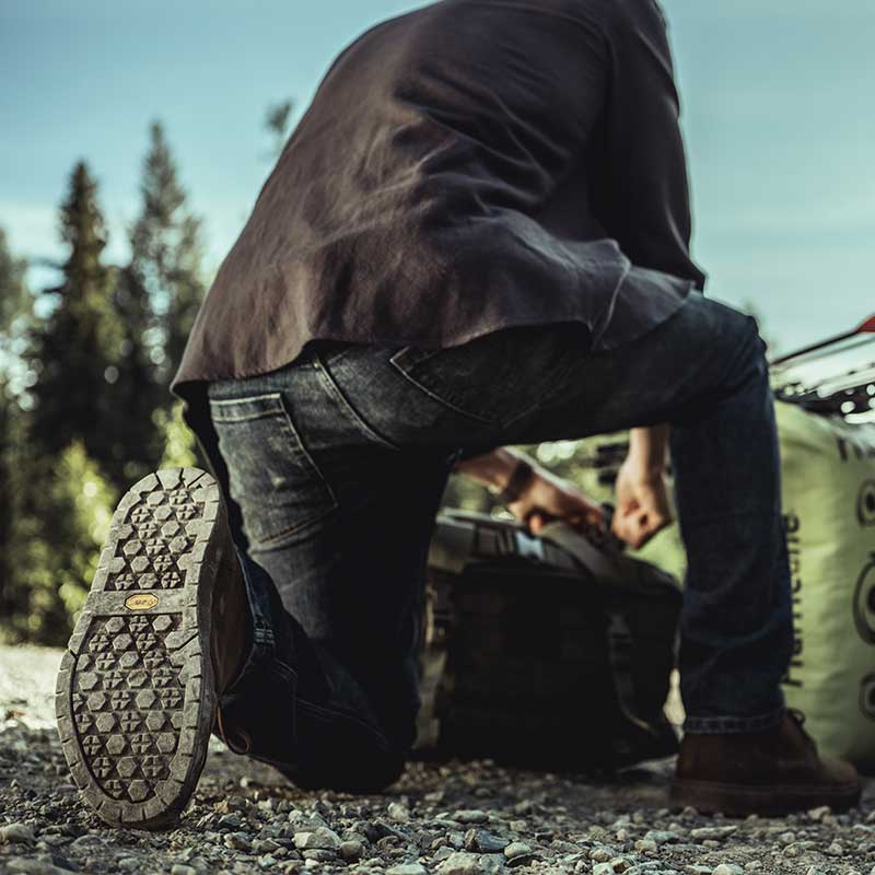 Ein Mann kniet, um etwas in seinem Rucksack zu verstellen, mit dem Rücken zur Kamera, was die Laufsohle eines Paares schwarzer Chukka-Stiefel zeigt.