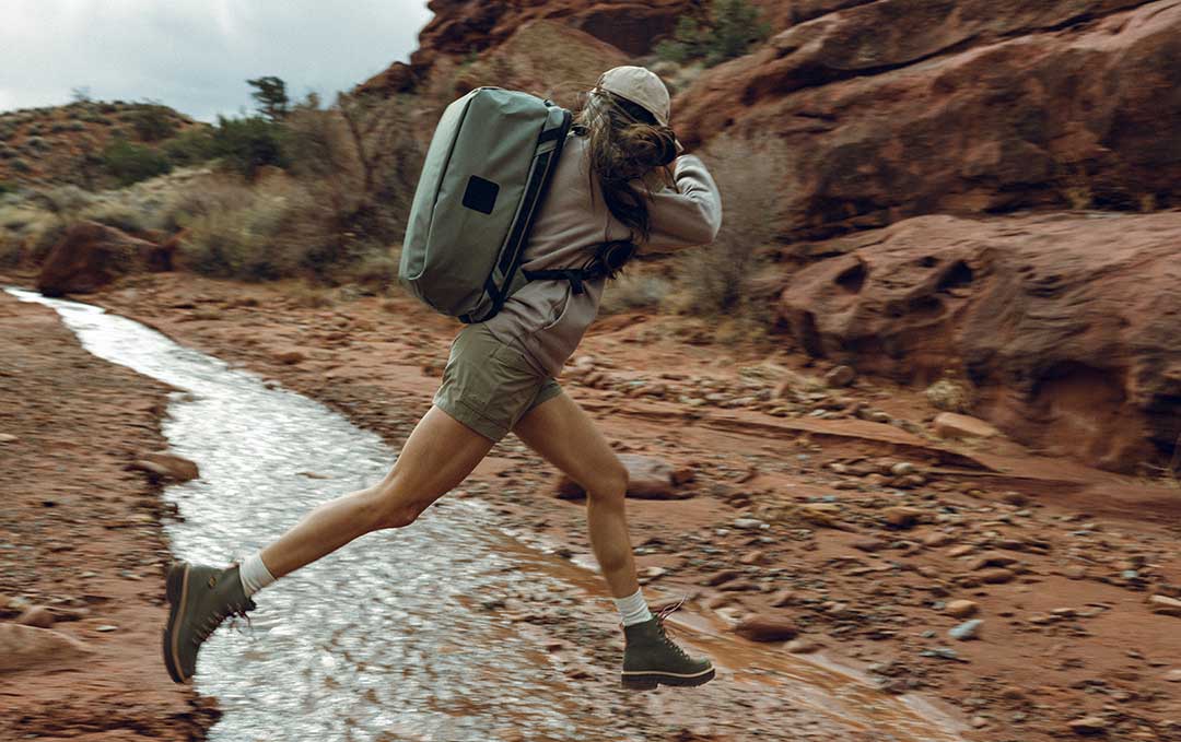 Woman carrying a light green duffel bag, jumping over a small creek.