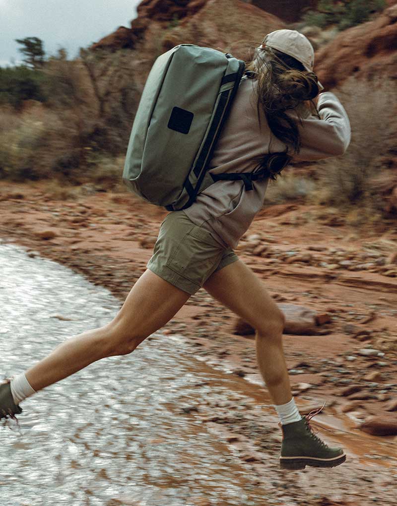 Woman carrying a light green duffel bag, jumping over a small creek.