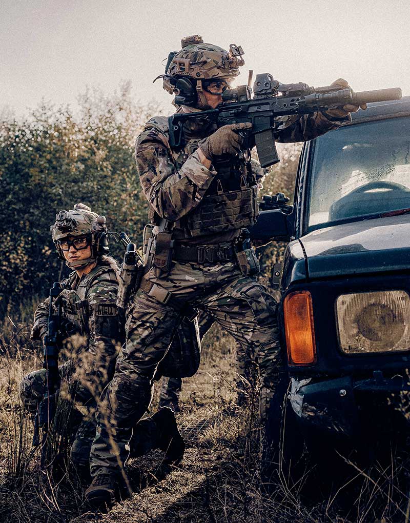Zwei Soldaten, die Tarnuniformen mit Cargo-Hosen tragen, sp&auml;hen hinter einem Auto hervor.