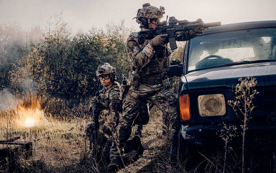 Zwei Soldaten, die Tarnuniformen mit Cargo-Hosen tragen, sp&auml;hen hinter einem Auto hervor.